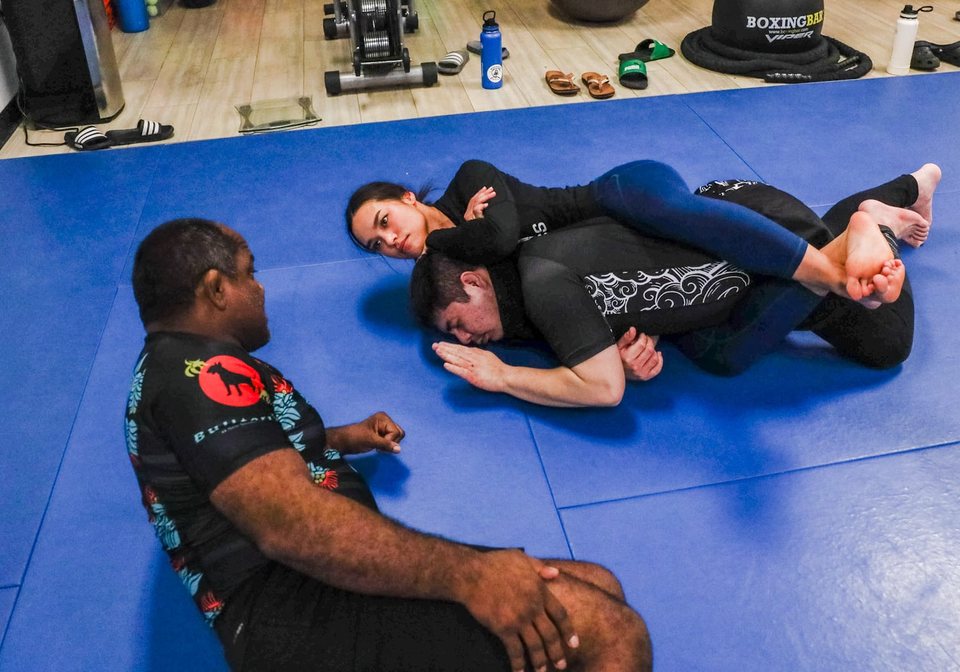 An instructor sits at the edge of the mat and watches closely as two students drill a position